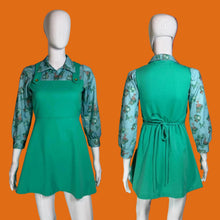 Load image into Gallery viewer, A vintage green pinafore dress displayed on a white mannequin, shown from both the front and back. The dress features a solid green pinafore bodice with button details and a tie waist, layered over a light blue floral long-sleeve shirt.