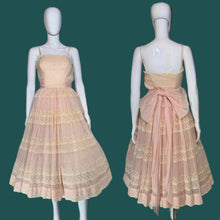 Load image into Gallery viewer, A sleeveless vintage dress displayed on a white mannequin from front and back views against a solid black background. The dress is made of a sheer, light peach-pink mesh with beige lace trim and features a large matching fabric bow at the back waist.