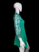 Load image into Gallery viewer, A green pinafore dress with three-quarter sleeves displayed on a white mannequin. The sleeves feature a light blue base with a whimsical cartoon character print in green, orange, and white.