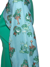 Load image into Gallery viewer, A close-up of a light blue fabric with a whimsical pattern of green animals, trees, and flowers, draped over a solid green garment against a neutral gray background.