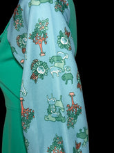 Load image into Gallery viewer, A close-up of a light blue fabric with a whimsical pattern of green animals, trees, and flowers, draped over a solid green garment against a neutral gray background.
