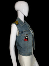 Load image into Gallery viewer, A light blue denim vest displayed on a white mannequin. The vest has yellow embroidered shoulder patches, a front snap closure, flap pockets, and a small red-and-black patch on the chest.