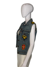 Load image into Gallery viewer, A light blue denim vest displayed on a white mannequin. The vest has military-style shoulder patches and two decorative embroidered patches on the front, one yellow eagle patch and one red and yellow shield patch.