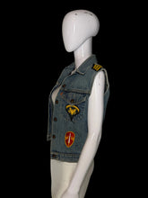 Load image into Gallery viewer, A light blue denim vest displayed on a white mannequin. The vest has military-style shoulder patches and two decorative embroidered patches on the front, one yellow eagle patch and one red and yellow shield patch.