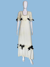 Load image into Gallery viewer, A vintage-style cream maxi dress displayed on a white mannequin, featuring black ribbon bows, lace trim, and a long black sash at the back.