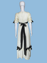 Load image into Gallery viewer, A vintage-style cream maxi dress displayed on a white mannequin from both front and back views, featuring black ribbon bows, lace trim, and a long black sash at the back.