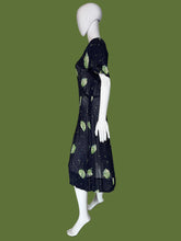 Load image into Gallery viewer, A sheer navy blue V-neck dress displayed on a white mannequin, featuring a green leaf print and small scattered cream, green, and teal speckles.