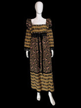 Load image into Gallery viewer, A long-sleeve maxi dress displayed on a white mannequin. The dress has a black base with yellow and orange horizontal stripes on the bodice and hem, and a central floral print panel in yellow, orange, and pink tones.