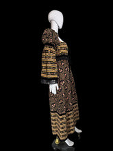 Load image into Gallery viewer, A long-sleeve maxi dress displayed on a white mannequin. The dress has a black base with yellow and orange horizontal stripes on the bodice and hem, and a central floral print panel in yellow, orange, and pink tones.