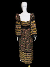 Load image into Gallery viewer, A back view of a long-sleeve maxi dress displayed on a white mannequin. The dress has a black base with yellow and orange horizontal stripes on the bodice and hem, and a central floral print panel in yellow, orange, and pink tones.