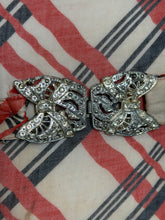 Load image into Gallery viewer, A 1930's silver bow belt buckle embellished with rhinestones, resting on a red, black, and cream plaid fabric background.