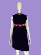 Load image into Gallery viewer, A sleeveless deep purple velvet dress displayed on a white mannequin. The dress features a high mock neck, a colorful patterned waistband with yellow, red, and blue floral motifs, and a matching patterned collar.