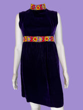 Load image into Gallery viewer, A sleeveless deep purple velvet dress displayed on a white mannequin. The dress features a high mock neck, a colorful patterned waistband with yellow, red, and blue floral motifs, and a matching patterned collar.