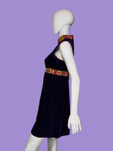 Load image into Gallery viewer, A sleeveless deep purple velvet dress displayed on a white mannequin. The dress features a high mock neck, a colorful patterned waistband with yellow, red, and blue floral motifs, and a matching patterned collar.