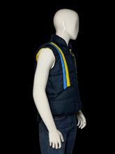Load image into Gallery viewer, A sleeveless navy quilted puffer vest with yellow and blue diagonal stripes on the chest, displayed on a white mannequin wearing blue jeans.