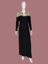Load image into Gallery viewer, A long black dress displayed on a white mannequin. The dress features a V-neckline with beige and gold patterned lace-like shoulder panels and long sleeves.