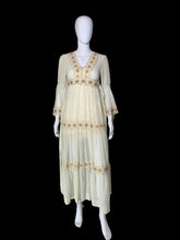 Load image into Gallery viewer, A cream-colored maxi dress with beige embroidery, shown on a white mannequin. The dress features a V-neckline, long bell sleeves, a tie waist, and tiered ruffles decorated with embroidered patterns.