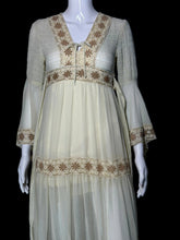 Load image into Gallery viewer, A close up of a cream-colored maxi dress with beige embroidery, shown on a white mannequin. The dress features a V-neckline, long bell sleeves, a tie waist, and tiered ruffles decorated with embroidered patterns.