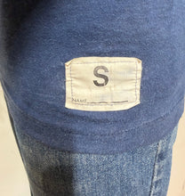 Load image into Gallery viewer, Close-up of a blue knit garment with a small beige fabric label stitched onto the side, showing a large letter S and the partial text 'NAME_______'.