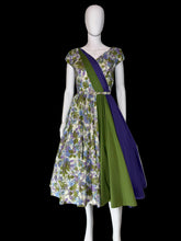 Load image into Gallery viewer, A vintage-style A-line dress displayed on a white mannequin. The dress features a floral print on one half and solid panels of green and purple on the other, with a matching waist belt.