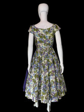 Load image into Gallery viewer, A short sleeve wrap-style dress displayed on a white mannequin, featuring a floral print bodice and skirt in cream, green, and purple, with a solid green and purple draped sash detail across the front.