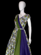 Load image into Gallery viewer, A short sleeve wrap-style dress displayed on a white mannequin, featuring a floral print bodice and skirt in cream, green, and purple, with a solid green and purple draped sash detail across the front.