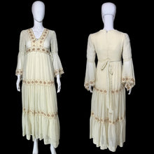 Load image into Gallery viewer, A cream-colored maxi dress with beige embroidery, shown from both the front and back on a white mannequin. The dress features a V-neckline, long bell sleeves, a tie waist, and tiered ruffles decorated with embroidered patterns.
