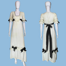 Load image into Gallery viewer, A vintage-style cream maxi dress displayed on a white mannequin from both front and back views, featuring black ribbon bows, lace trim, and a long black sash at the back.
