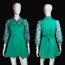 Load image into Gallery viewer, A vintage green pinafore dress displayed on a white mannequin, shown from both the front and back. The dress features a solid green pinafore bodice with button details and a tie waist, layered over a light blue floral long-sleeve shirt.
