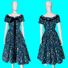 Load image into Gallery viewer, A vintage 1950s off-the-shoulder A-line dress shown on a white mannequin from both front and back views. The dress features a vibrant blue, purple, and green abstract pattern with black trim and a black bow at the neckline.
