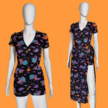 Load image into Gallery viewer, A women's short-sleeve romper and matching wrap skirt set displayed on white mannequins against a plain indoor wall. The set features a black base with a colorful abstract cloud-like print in purple, teal, orange, and pink.
