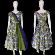Load image into Gallery viewer, A vintage-style floral dress displayed on a white mannequin, shown from the front and back. The dress has a multicolor floral print with green, purple, blue, and white tones, plus a solid green draped sash and a dark purple side panel.
