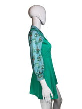 Load image into Gallery viewer, A green pinafore dress with three-quarter sleeves displayed on a white mannequin. The sleeves feature a light blue base with a whimsical cartoon character print in green, orange, and white.

