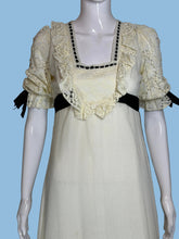 Load image into Gallery viewer, A vintage-style cream maxi dress displayed on a white mannequin from both front and back views, featuring black ribbon bows, lace trim, and a long black sash at the back.
