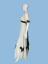 Load image into Gallery viewer, A vintage-style cream maxi dress displayed on a white mannequin from both front and back views, featuring black ribbon bows, lace trim, and a long black sash at the back.

