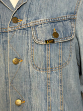 Load image into Gallery viewer, Close-up of a light blue denim jacket with gold-tone Lee branded buttons and a visible Lee brand patch on the chest pocket.
