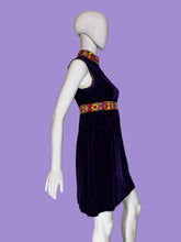 Load image into Gallery viewer, A sleeveless deep purple velvet dress displayed on a white mannequin. The dress features a high mock neck, a colorful patterned waistband with yellow, red, and blue floral motifs, and a matching patterned collar.
