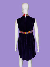 Load image into Gallery viewer, A sleeveless deep purple velvet dress displayed on a white mannequin. The dress features a high mock neck, a colorful patterned waistband with yellow, red, and blue floral motifs, and a matching patterned collar.
