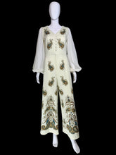 Load image into Gallery viewer, A cream-colored jumpsuit with sheer long sleeves and ornate paisley embroidery in gold, green, and blue, displayed on a white mannequin.
