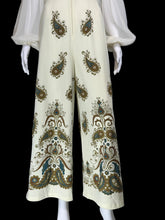 Load image into Gallery viewer, A shot of the bottom half of the jumpsuit displayed on a mannequin. The pants are cream-colored with a prominent paisley pattern in brown, teal, and gold.

