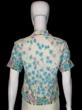 Load image into Gallery viewer, A vintage short-sleeve collared blouse with a blue, pink, and green floral print on an off-white background, displayed on a white mannequin from the back.
