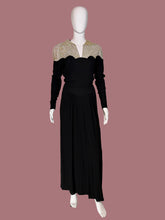 Load image into Gallery viewer, A long black dress displayed on a white mannequin. The dress features a V-neckline with beige and gold patterned lace-like shoulder panels and long sleeves.
