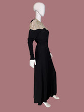 Load image into Gallery viewer, A long black dress displayed on a white mannequin from the side. The dress features a V-neckline with beige and gold patterned lace-like shoulder panels and long sleeves.

