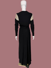 Load image into Gallery viewer, A long black dress displayed on a white mannequin from the back. The dress features a V-neckline with beige and gold patterned lace-like shoulder panels and long sleeves.
