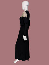 Load image into Gallery viewer, A long black dress displayed on a white mannequin from the side. The dress features a V-neckline with beige and gold patterned lace-like shoulder panels and long sleeves.
