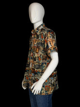 Load image into Gallery viewer, A short-sleeve button-up shirt displayed on a white mannequin. The shirt features a dense collage print of magazine cover illustrations in a multicolor palette of blue, red, beige, and brown.
