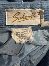 Load image into Gallery viewer, Close-up of a blue denim garment showing a sewn-on Sakowits brand label and a torn interior size tag.
