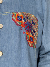 Load image into Gallery viewer, Close-up of a blue denim shirt featuring a large, colorful embroidered patch on the chest with geometric patterns in red, orange, green, and blue.
