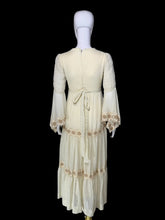 Load image into Gallery viewer, A back view of a cream-colored maxi dress with beige embroidery, shown on a white mannequin. The dress features a V-neckline, long bell sleeves, a tie waist, and tiered ruffles decorated with embroidered patterns.
