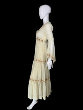 Load image into Gallery viewer, A side view of a cream-colored maxi dress with beige embroidery, shown on a white mannequin. The dress features a V-neckline, long bell sleeves, a tie waist, and tiered ruffles decorated with embroidered patterns.
